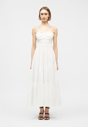 Vrouw draagt een witte maxi-jurk met meerdere lagen en dunne bandjes, gecombineerd met zwarte sandalen met hoge hakken en bandjes, staand tegen een effen lichte achtergrond.