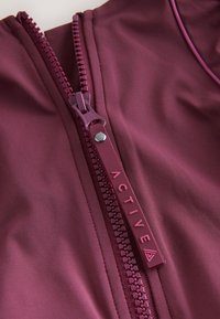 Gros plan d'une fermeture éclair ouverte d'une veste violette avec une languette portant l'inscription "ACTIVE" et un petit logo triangulaire en bas.