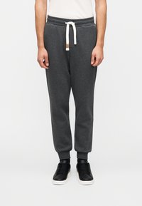 Σκούρα γκρι φόρμες με κορδόνι στη μέση, ριμπ γιακάδες και μια μικρή δερμάτινη ετικέτα στο μπροστινό μέρος. Συνδυάζονται με μαύρα αθλητικά παπούτσια.