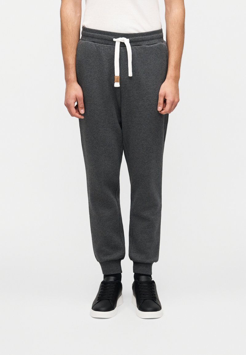 Σκούρα γκρι φόρμες με κορδόνι στη μέση, ριμπ γιακάδες και μια μικρή δερμάτινη ετικέτα στο μπροστινό μέρος. Συνδυάζονται με μαύρα αθλητικά παπούτσια.