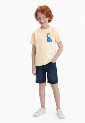 Gutt med rødt hår smiler, iført lys oransje t-skjorte med blå dinosaur og teksten "RAWR!", mørke denimshorts og hvite joggesko.