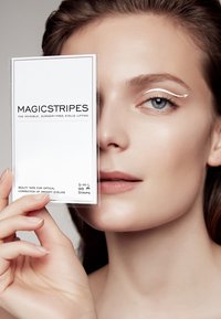 Packaging del nastro sollevante per palpebre Magicstripes tenuto da una persona. Presenta uno sfondo bianco con testo nero e una finestra trasparente. 96 strisce, taglie S, M, L.
