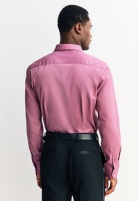 Dopasowana koszula z długim rękawem w jasnoróżowym materiale, z kołnierzykiem i mankietami na guziki, zestawiona z czarnymi spodniami i czarnym paskiem.