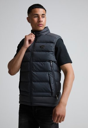 Jonge man draagt een zwart gewatteerd vest over een zwart T-shirt, ritst deze dicht, staand voor een effen lichtgrijze achtergrond.