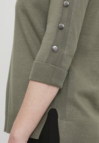 Olīvu zaļš džemperis ar mīkstu tekstūru, ar ribinātu aproci un pieciem metāliskiem pogām pa piedurkni. Norādītas sānu šķiršanās detaļas.