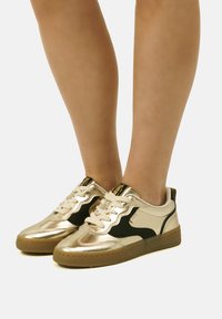 Zapatillas metálicas doradas con acentos negros, suela de goma texturizada, diseño de cordones y cuello acolchado. Elegantes y modernas con colores contrastantes.