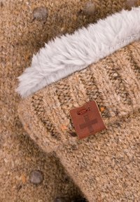 Knitted wool beanie in tan with a soft grey faux fur lining. Features a brown leather tag with a cross emblem and colorful specks throughout.