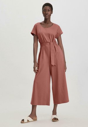 Combinaison couleur rouille à manches courtes, design à jambes larges, taille cintrée avec un noeud, fabriquée en tissu lisse. Portée avec des sandales légères.