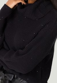 Primer plano de una persona que lleva un suéter negro de cuello alto con pequeñas lentejuelas brillantes y pantalones de cuero negros con un cinturón de lazo.