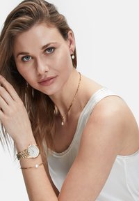 Guldfarvet ure med hvid urskive, parret med et guldkædearmbånd med en perleaccent. Modellen bærer også en guldkædesmykke med en perle.