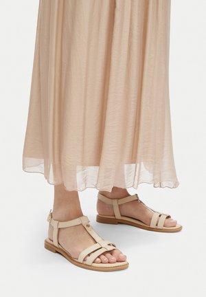 Béžové sandály s otevřenou špičkou a T-proužkem. Plochá podrážka s tenkým páskem kolem kotníku a zadní sponou. Kombinováno s lehkou, béžovou sukní s volným střihem.