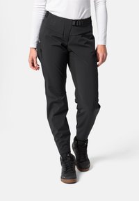 Persoon in zwarte buitenbroek met riem en zwarte wandelschoenen, gecombineerd met een witte lange mouwen top, staand tegen een witte achtergrond.