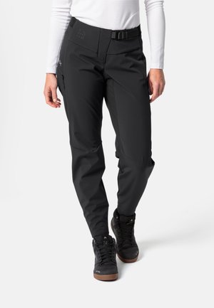 Persoon in zwarte buitenbroek met riem en zwarte wandelschoenen, gecombineerd met een witte lange mouwen top, staand tegen een witte achtergrond.