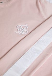 Helles rosa Sweatshirt mit weißem, aufwendig gesticktem Logo auf der Brust und einem weißen Stoffstreifen mit dezentem Text am Ärmel.