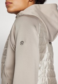 Giacca beige con cappuccio, dotata di un pannello laterale trapuntato di colore argento e un logo metallico rotondo sulla manica. La texture morbida si contrappone al tessuto liscio.