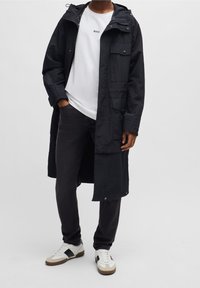 Dlouhá černá parka s kapucí, několika kapsami a zipem, nošená přes bílé tričko a tmavé džíny, doplněná bílými teniskami.