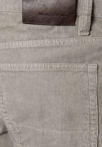 Beżowy sztruks z pionowymi prążkami, wyposażony w tylną kieszeń oraz ciemnobrązową skórzaną etykietę z wytłoczonym napisem „Marc O'Polo”.