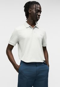 Ανοικτό γκρι πόλο με γιακά και κοντά μανίκια, με διακριτικές ριμπ λεπτομέρειες και λογότυπο στο στήθος, συνδυασμένο με σκουρό μπλε παντελόνι.