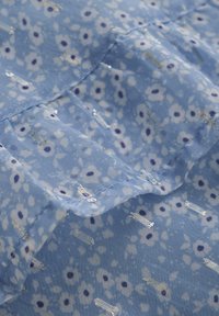 Lichtblauwe stof met een bloempatroon bestaande uit witte en donkerblauwe bloemen en zilveren metallic accenten; licht gekreukelde textuur.