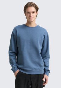 Niebieska bluza z długim rękawem, okrągłym dekoltem oraz prążkowanymi mankietami i dołem. Gładki materiał o luźnym kroju. Noszona z czarnymi spodniami.