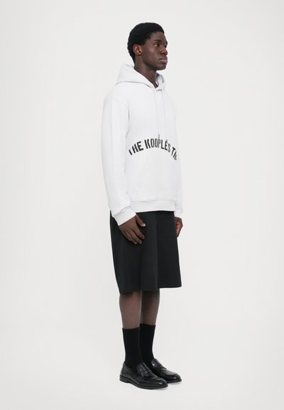 Junger Mann steht in einem weißen Hoodie mit Aufschrift, schwarzen knielangen Shorts, schwarzen Socken und schwarzen Lederloafers vor einem einfarbigen Hintergrund.