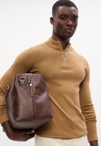 Borsa in pelle marrone con una forma strutturata, dotata di chiusura con zip e dettagli in metallo laterali. Il modello indossa un top lungo in tonalità beige con zip sul collo.