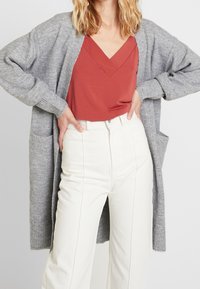 Grå stickad cardigan med ribbad ärmslut och fickor, lager på lager över en rostfärgad v-ringad topp och kombinerad med vita högmidjade byxor.