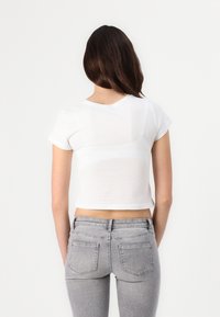 Camiseta blanca corta de manga corta con escote redondo, mostrada desde la parte de atrás. Combinada con jeans ajustados de color gris claro. Textura de tela suave.