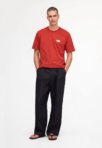 Man stands with hands in pockets wearing a red short-sleeve T-shirt with logo, loose black pants, and black flip-flops on a white background.