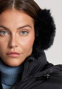 Cuffie nere con bordo in pelliccia sintetica, indossate con un maglione a collo alto blu in maglia sotto una giacca nera con zip. Pelle liscia e trucco neutro.