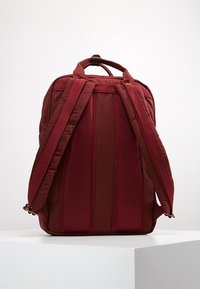 Czerwona plecak z poduszkowanymi siateczkowymi paskami, usztywnioną konstrukcją i gładkim wykończeniem materiału; posiada zamki błyskawiczne i akcenty z złotego metalu.