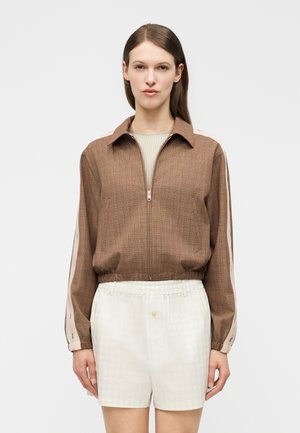 Femme portant une veste marron zippée avec des rayures beige clair sur les manches et un short blanc taille haute sur un fond uni.