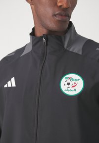 Chaqueta negra con cuello alto, que cuenta con un cierre frontal, acentos grises y un logo del equipo nacional de Argelia con un balón de fútbol y estrellas.