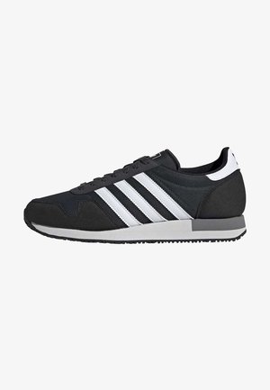 Baskets Adidas basses noires et blanches avec trois bandes blanches, lacets noirs et semelle grise et blanche.