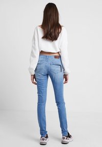 Hellblaue Skinny-Jeans mit einer mittelhohen Taille, ausgestattet mit Gesäßtaschen und einer strukturierten Oberfläche. Kombiniert mit einem weißen, cropped Pullover.