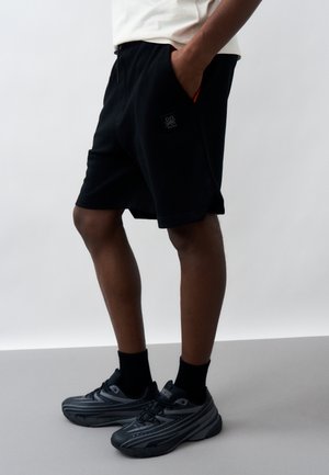 Mann trägt schwarze Shorts, schwarze Socken und graue Sneakers, steht mit einer Hand in der Tasche vor einem einfarbigen Hintergrund.