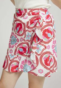 Jupe portefeuille florale blanche avec des motifs rouges, roses et bleus, dotée d'un bord volanté et d'un lien ajustable à la taille pour un look ajusté.