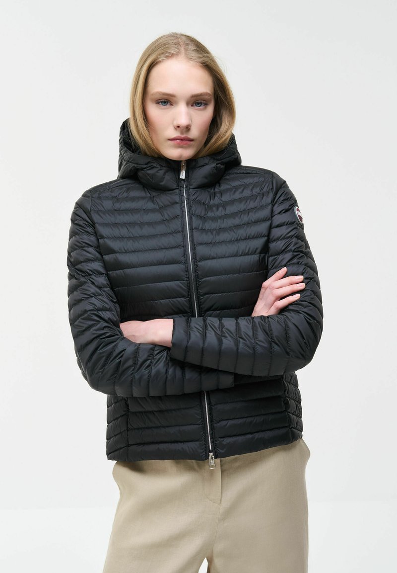 Mladá žena se světlými vlasy v černé prošívané bundě s kapucí a béžových kalhotách, stojící s překříženýma rukama na bílém pozadí.