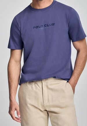 Mężczyzna w niebieskiej koszulce "Polo Club" i beżowych spodniach, z jedną ręką w kieszeni, a drugą luźno opuszczoną wzdłuż ciała.