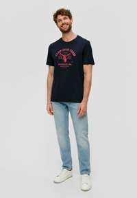 Tmavě modré bavlněné tričko s grafikou "CLIFF DIVE TEAM" v růžové, kulatým výstřihem, krátkými rukávy, nosí se s světle modrými džíny a bílými teniskami.