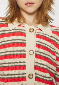 Cardigan lavorato a maglia con strisce rosse, beige e grigie. Presenta una chiusura frontale con bottoni, colletto e tessuto testurizzato. Bottoni grandi notevoli.