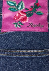 Jeans de mezclilla con una etiqueta de satén rosa que presenta rosas rojas bordadas y hojas verdes, acentuadas por la palabra "Replay" en negro.