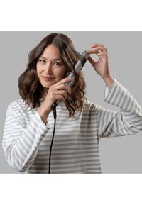 Dispositivo per arricciare i capelli con un cilindro grigio e una maniglia testurizzata; l'utente dimostra come arricciare i capelli. L'utente indossa una maglietta a righe grigie e bianche.