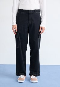 Black cargo pants with wide legs, contrast stitching, two side pockets, and a high waistband, paired with brown sneakers and white laces.