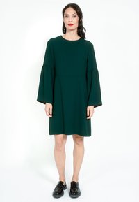 Zelena haljina s dugim zvonastim rukavima, okruglim izrezom i slobodnim krojem. Glatka tekstura s jednostavnim dizajnom, uparena s crnim loafericama.