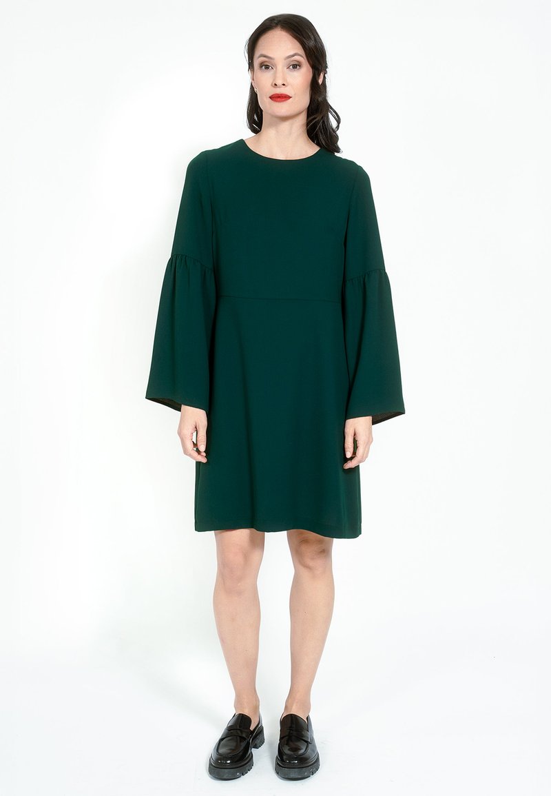 Zelena haljina s dugim zvonastim rukavima, okruglim izrezom i slobodnim krojem. Glatka tekstura s jednostavnim dizajnom, uparena s crnim loafericama.