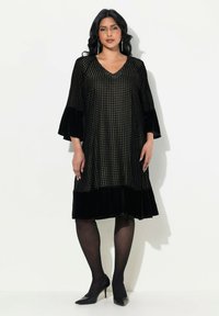 Schwarzes Kleid mit V-Ausschnitt, strukturiertem Stoff mit einem Hahnentritt-Muster, Samtärmeln und einem ausgestellten Rock. Getragen mit schwarzen Absätzen.