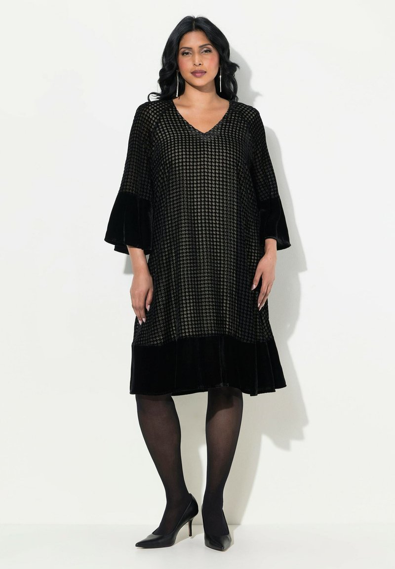 Schwarzes Kleid mit V-Ausschnitt, strukturiertem Stoff mit einem Hahnentritt-Muster, Samtärmeln und einem ausgestellten Rock. Getragen mit schwarzen Absätzen.
