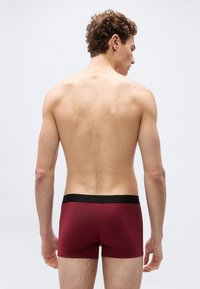 Νεαρός άνδρας με σγουρά μαλλιά, γυρισμένος με την πλάτη στην κάμερα, φοράει σκούρα κόκκινα αντρικά εσώρουχα μποξέρ με μαύρη μέση, εναντίον απλού φόντου.