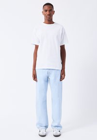 Hvid bomulds T-shirt med korte ærmer, parret med lyseblå lige-lår jeans. Modellen bærer grå og hvide sneakers. Ensfarvet baggrund.